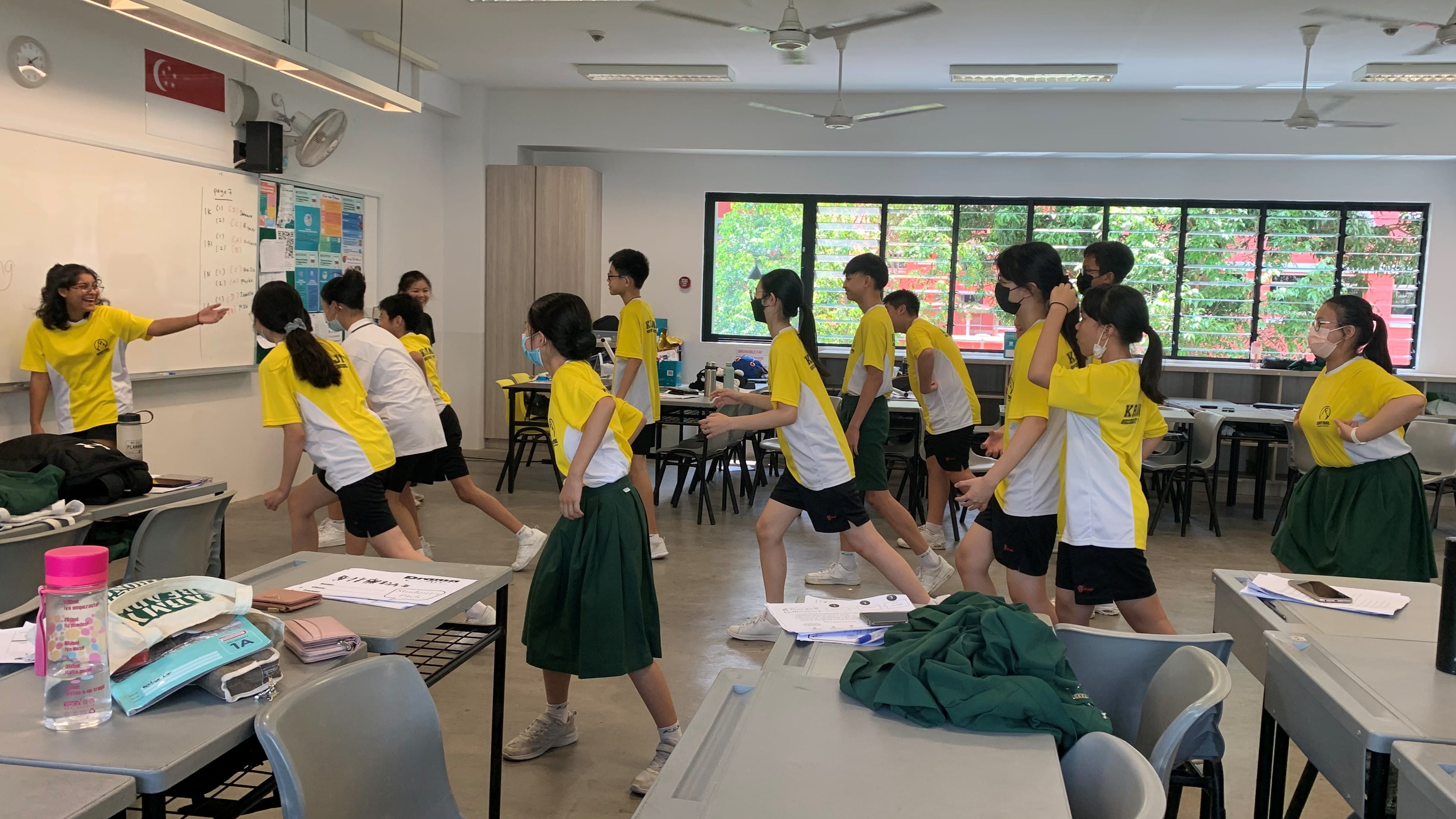 Students in yellow uniforms doing physical activity in a classroom while a teacher gestures near a whiteboard.